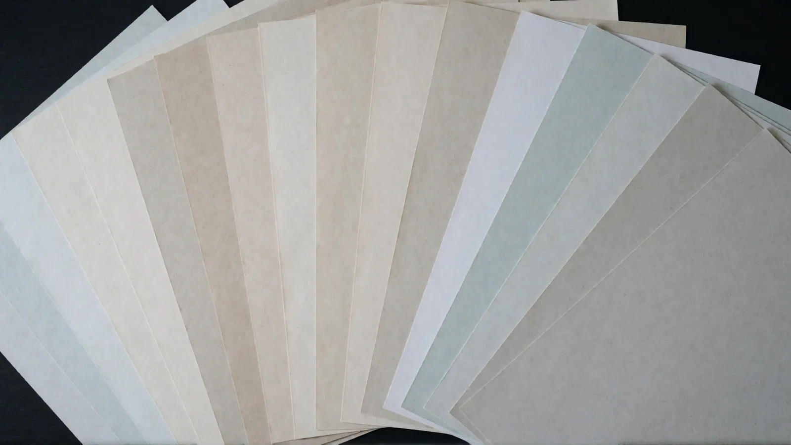 Sheets of blank paper in twenty-four different tints laid out in a grid on an oak desk, with no writing, no marking and no printing on any of them